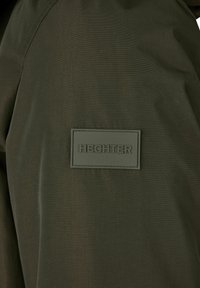 Donkergroene stof met een gladde textuur heeft een rechthoekige rubberen patch met de opdruk "HECHTER." Close-up van het kledingstuk.