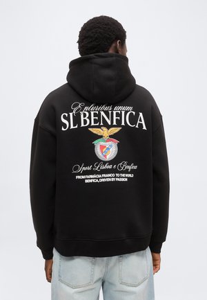 Άτομο που φοράει μαύρο φούτερ με κουκούλα με το λογότυπο του SL Benfica και το κείμενο «E pluribus unum» και «From Farmácia Franco to the world» στην πλάτη.