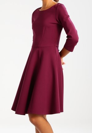 Femme portant une robe bordeaux foncé longueur genou, avec des manches trois-quarts et une jupe évasée, debout les mains derrière le dos.