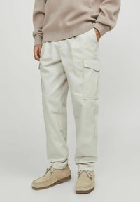 Pantaloni cargo beige con tasche laterali, vestibilità sartoriale, materiale in cotone e vita elastica. Indossati con scarpe in suede beige chiaro.
