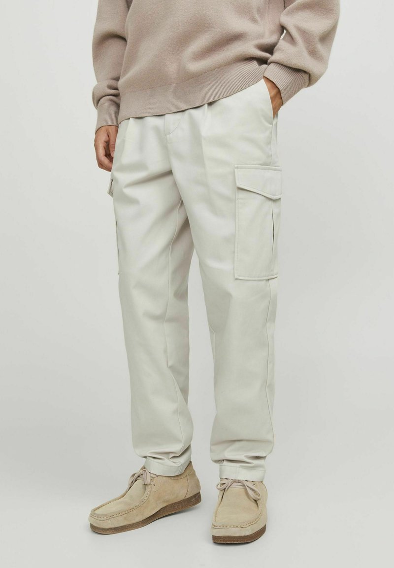 Pantaloni cargo beige con tasche laterali, vestibilità sartoriale, materiale in cotone e vita elastica. Indossati con scarpe in suede beige chiaro.