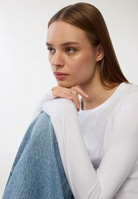 Maglia bianca a maniche lunghe e tessuto denim blu chiaro, che presenta una vestibilità rilassata. Texture liscia con design semplice e dettagli minimi.