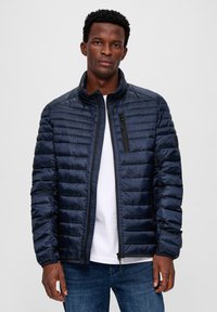 Man in een donkerblauwe gewatteerde jas over een wit shirt en blauwe jeans, staande tegen een effen lichte achtergrond.