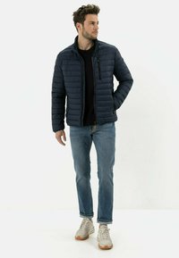 Marineblaue gepolsterte Jacke mit vertikaler Steppoptik, hohem Kragen und Reißverschluss. Getragen über einem schwarzen Hemd, kombiniert mit hellen Jeans und Turnschuhen.