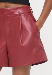 Pantaloni corti in finta pelle bordeaux con vita alta, tasche laterali e pieghe. Il tessuto ha una consistenza liscia e lucida.