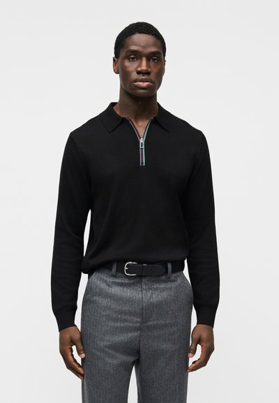 Pull polo noir à manches longues avec col zippé et bordures colorées. Associé à un pantalon gris à fines rayures et une ceinture noire.