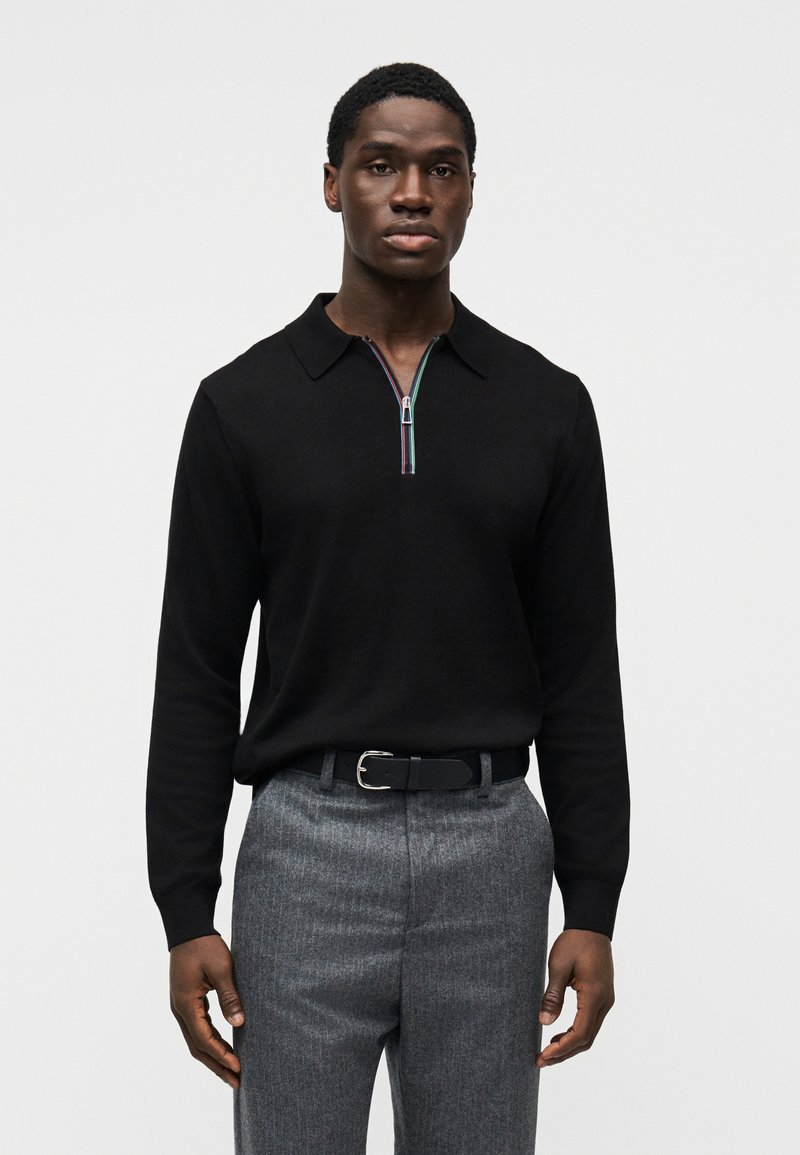 Μαύρο πλεκτό πόλο με μακριά μανίκια και φερμουάρ στο λαιμό, με πολύχρωμες λεπτομέρειες. Συνδυάζεται με γκρι παντελόνια με ρίγες και μαύρη ζώνη.