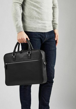 Sort læder arbejdsmappe med dobbelte topgreb, sølvfarvet beslag og en lynlåslomme foran, holdt af en person iført en lysegrå sweater.
