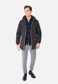 Giacca a vento nera in nylon con cappuccio, chiusura con zip e dettagli in rosa oro a contrasto. Abbinata a un maglione blu a lavorazione a trecce e pantaloni grigi.