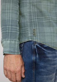 Camisa de cuadros verde hecha de tela texturizada. Presenta un dobladillo curvado y una etiqueta con números en la parte inferior. Combinada con vaqueros de mezclilla azul.