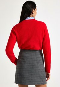 Kvinna med långt mörkt hår, iförd röd tröja, blå blus med volangkrage, guldörhängen och grå rutad kjol, stående med handen på höften.