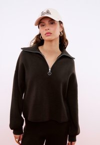 Übergroßer dunkelgrüner Pullover mit einem halben Reißverschluss am Kragen und Rippbündchen, kombiniert mit einer cremefarbenen Mütze mit dem Schriftzug "LA" in lässiger Schriftart.