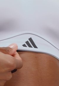 Bijela sportska grudnjak s crnim Adidas logom i zakrivljenim dizajnom. Tkanina izgleda glatko s uskim fitom, ističući kožu ispod.