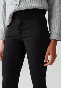 Persoon draagt strakke zwarte jeans en een grijze geribbelde gebreide trui met opgerolde mouwen bij de polsen, staand tegen een effen achtergrond.
