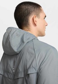 Giacca con cappuccio grigio chiaro dal texture liscia e leggermente riflettente. Presenta una aletta posteriore e accenti riflettenti sottili sulla parte posteriore.