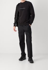 Czarna bluza z logo "Calvin Klein Jeans" z przodu, w połączeniu z czarnymi spodniami typu cargo i czarnymi butami na sznurowadła. Niosąc małą czarną torbę.