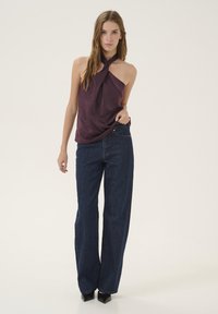 Donkerpaarse mouwloze top met een geknoopte halsdetail, gecombineerd met high-waisted, wijde donkerblauwe jeans en zwarte hakken.