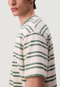 Camiseta a rayas en color crema con rayas verdes, de mangas cortas, con cuello de canalé y textura suave. Presenta un corte holgado y un diseño sencillo.