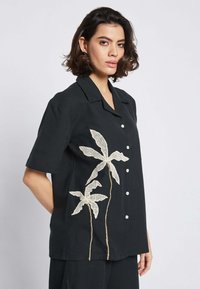 Camicia nera a maniche corte con ricami floreali bianchi, dotata di collarino e chiusura frontale con bottoni. Tessuto testurizzato con una vestibilità rilassata.