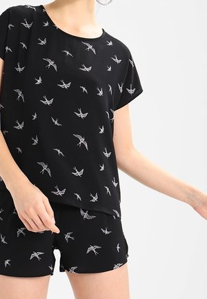 Kobieta ubrana w czarny zestaw z krótkim rękawem i szorty z białym nadrukiem ptaka na jednolitym jasnym tle.