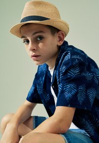 Blaue Kurzarmbluse mit Blattmuster, getragen mit einem weißen T-Shirt und Jeansshorts. Strohhut mit marineblauem Band. Person sitzt und schaut in die Kamera.