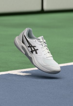 Fehér és szürke Asics teniszcipő fekete logóval, amely lebeg a kék teniszpálya felülete felett, közel a fehér határvonalhoz.