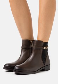 Bottines en cuir marron avec un panneau latéral élastique noir et des détails en métal doré. Présentent un bout arrondi et un petit talon carré.