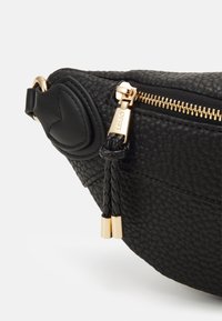 Schwarze Lederhandtasche mit strukturiertem Finish, goldfarbigem Reißverschluss, geflochtenem Detail und einem herzförmigen Akzent an der Seite.