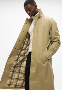 Beige overcoat met een effen buitenkant en een geruite binnenkant, met een hoge kraag en verborgen knoopsluiting.