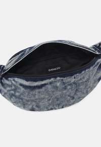 Tumma sininen denimvyölaukku, jossa on teksturoitu pinta, vetoketjusuljenta ja musta sisävuori, jossa merkintä näkyvästi esillä.