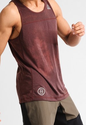 Muskulöse Person, die ein ärmelloses, kastanienfarbenes Reebok-Tanktop und zweifarbige Shorts in Beige und Schwarz trägt, in einer dynamischen Pose vor einem schlichten Hintergrund.