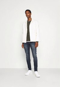 Chaqueta blanca sobre una camisa verde oliva, combinada con jeans azul oscuro y zapatillas blancas. El conjunto presenta un ajuste relajado y un estilo casual.