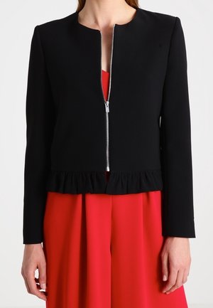 Femme portant une veste courte noire à manches longues avec une fermeture éclair devant sur une robe rouge plissée.