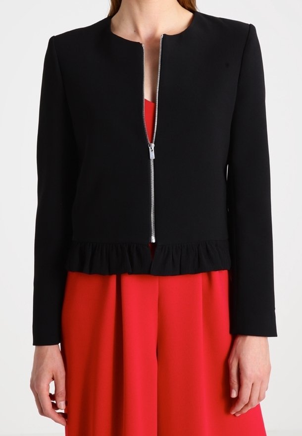Femme portant une veste courte noire à manches longues avec une fermeture éclair devant sur une robe rouge plissée.