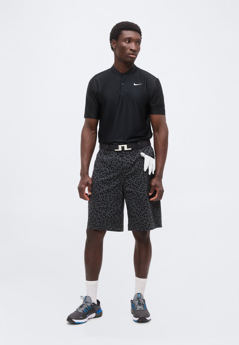 Homme portant un polo noir Nike, un short imprimé léopard noir et gris, un gant de golf blanc glissé dans le short, des chaussettes blanches et des baskets noires.