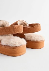 Brune suedestrop sandal med platform og fluffy beige pelsindlæg samt justerbare remme. Tekstureret overflade og forhøjet bund for komfort.