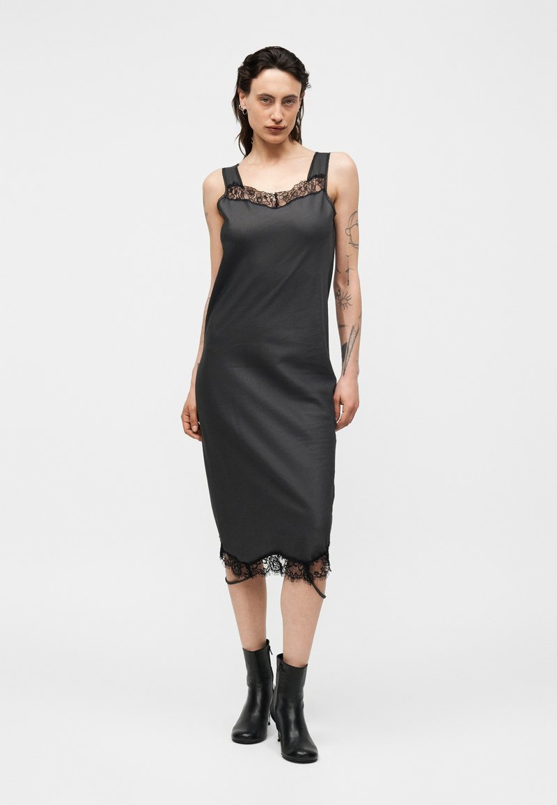 Frau mit Tattoos, die ein schwarzes Satin-Midikleid mit Spitzenbesatz und schwarze Knöchelstiefel trägt, steht vor einem schlichten hellen Hintergrund.