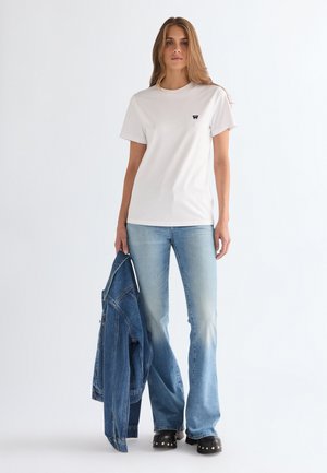 Γυναίκα που φοράει άσπρο κοντομάνικο μπλουζάκι και ανοιχτόμπλε τζιν με καμπάνα, κρατώντας ένα μπλε τζιν μπουφάν στο αριστερό της χέρι, στέκεται μπροστά σε απλό λευκό φόντο.