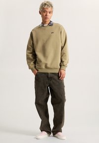 Sudadera de color verde oliva claro con cuello redondo, cuello decorado en gris, y vaqueros negros de corte holgado. Zapatillas blancas con detalles a cuadros en rosa.