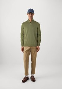 Zeleni rebrasti pulover z zadrgo do polovice in majhnim logotipom, kombiniran s bež hlačami in rjavimi loaferji. Model nosi mornarsko modro kapo in bele nogavice.