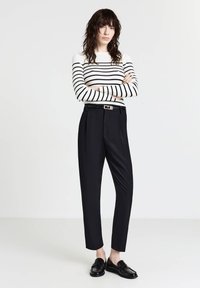 Pantalons noirs sur mesure avec des plis, associés à un pull rayé noir et blanc à côtes. Le modèle porte des mocassins noirs brillants.