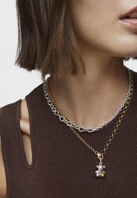 Dos collares en capas: una gruesa cadena plateada con eslabones ovalados y una cadena dorada con un pequeño colgante con acentos de perla, llevada sobre una blusa marrón.