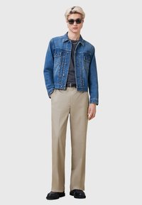 Denimjack in middelblauw, van katoen; grijze T-shirt eronder; beige broek; zwarte schoenen; zilveren kettinghalsband; zonnebril.