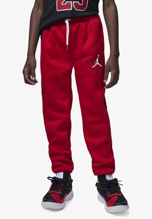 Pantalones de chándal rojos con cintura ajustable mediante cordón y detalles del logo en blanco. Combinados con zapatillas deportivas negras y rojas que tienen una suela texturizada.