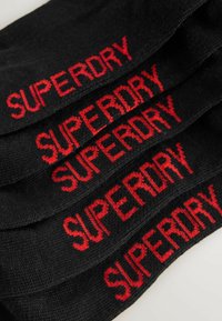 Juodos kojinės su raudonu "SUPERDRY" logotipu, sudėtos eilėmis. Medžiaga atrodo minkšta su megztu tekstūru. Logotipas turi ryškų, bloko tipo šriftą.