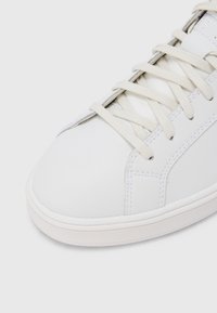 Λευκό δερμάτινο αθλητικό παπούτσι με στρογγυλεμένη μύτη, υφή στην επιφάνεια και κορδόνια σε κρεμ χρώ μα. Διαθέτει ραφές και λαστιχένια σόλα.