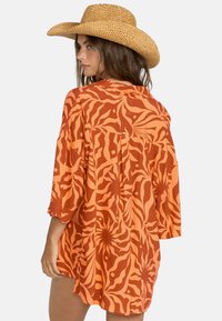 Camisa de patrón naranja con un corte holgado, que presenta formas en espiral en un naranja más oscuro, textura suave y un detalle de pliegue en la parte posterior.