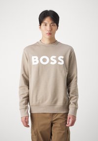 Sudadera beige con cuello redondo y puños acanalados. Presenta un gran texto blanco "BOSS" en la parte frontal. Textura simple y suave.