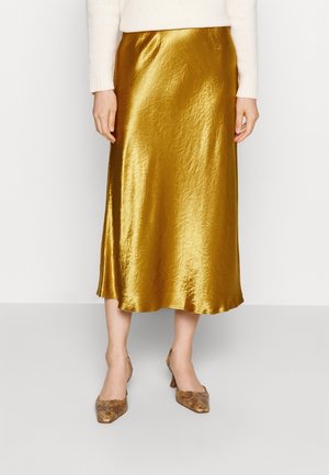 Vrouw die een glanzende gouden midirok draagt, een crèmekleurige trui en puntige hoge hakken met slangenprint, tegen een effen witte achtergrond.