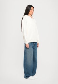 Lichtbeige hoodie met een voorzak en trekkoorden, gecombineerd met wijde blauwe jeans en witte sneakers. Glad stofmateriaal.